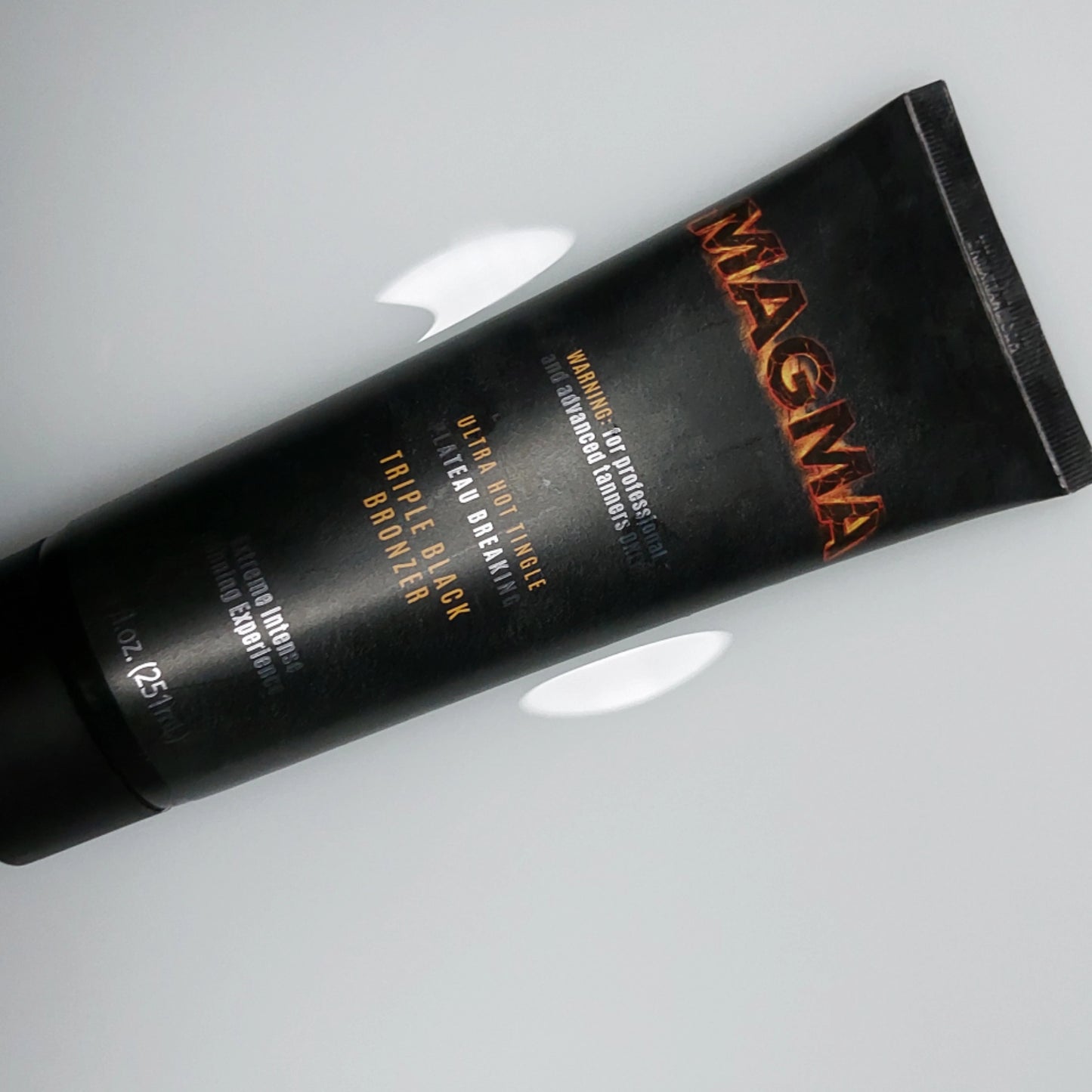 Die Onyx Magma Triple Black Bronzer Tube liegt flach auf einem weißen Hintergrund mit Cremeflecken, zeigt deutlich das Design und die Branding-Elemente der extrem heißen Tingle-Formel