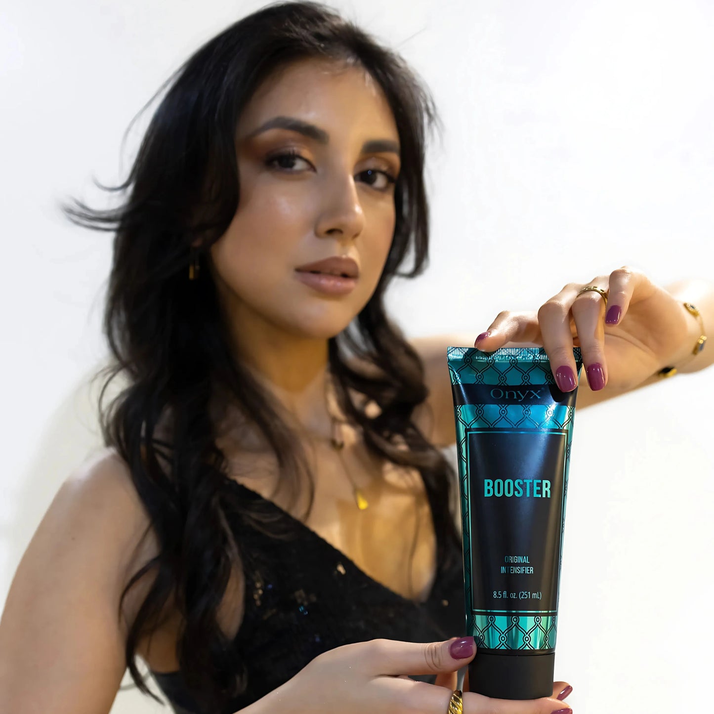 Frau im schwarzen Kleid präsentiert die Onyx Tanning Booster Bräunungslotion mit beiden Händen vor weißem Hintergrund – eleganter Beauty-Moment